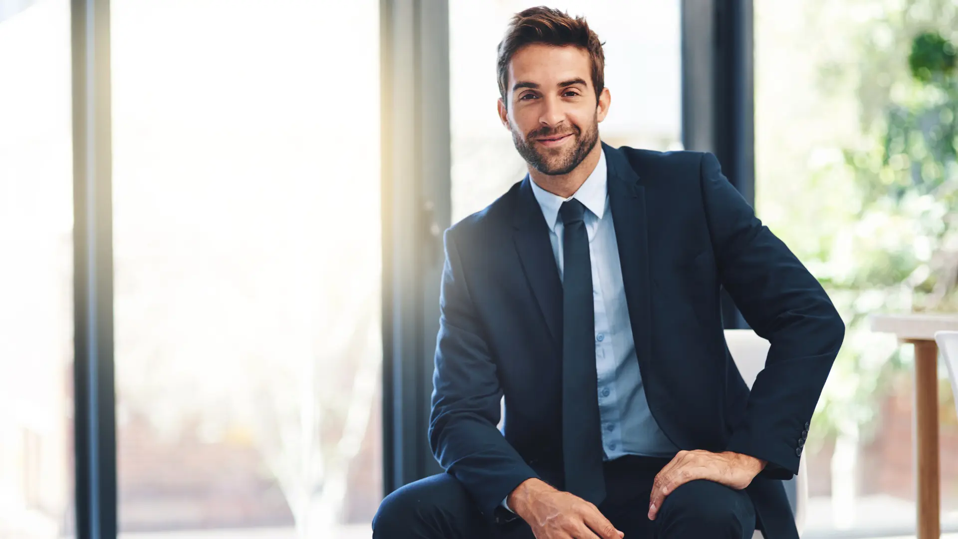 Un professionnel assis sur un bureau, détendu et souriant, une main sur la cuisse.